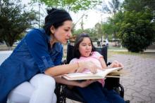 Image of parent with child with disabilities. They are outside and she is helping her with a book. 